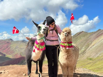 TourCompass-kollega Iida står med to flagg og en alpakka på toppen av Rainbow Mountain i Peru.